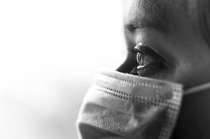 Close up black and white image depicting a caucasian woman, in her 30s with brown hair, wearing a blue surgical face mask for safety and protection during the coronavirus (Covid-19) pandemic. The woman has intense brown eyes which are in sharp focus. Room for copy space.