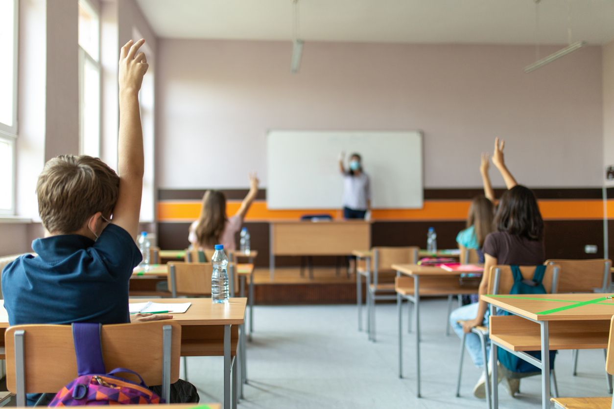 Students with protective masks sitting in school desks in their classroom. School desks are marked with a cross to mark a place where sitting is not allowed to maintain social distance during corona virus .