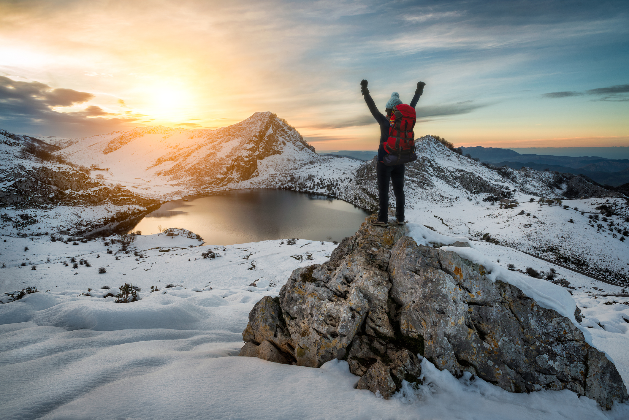 Hiker woman with backpack and hat rising arms in victory sign on snowy mountain peak at Covadonga lake, beautiful snow winter landscape sunset scene on a touristic location of Asturias, Spain, Europe.