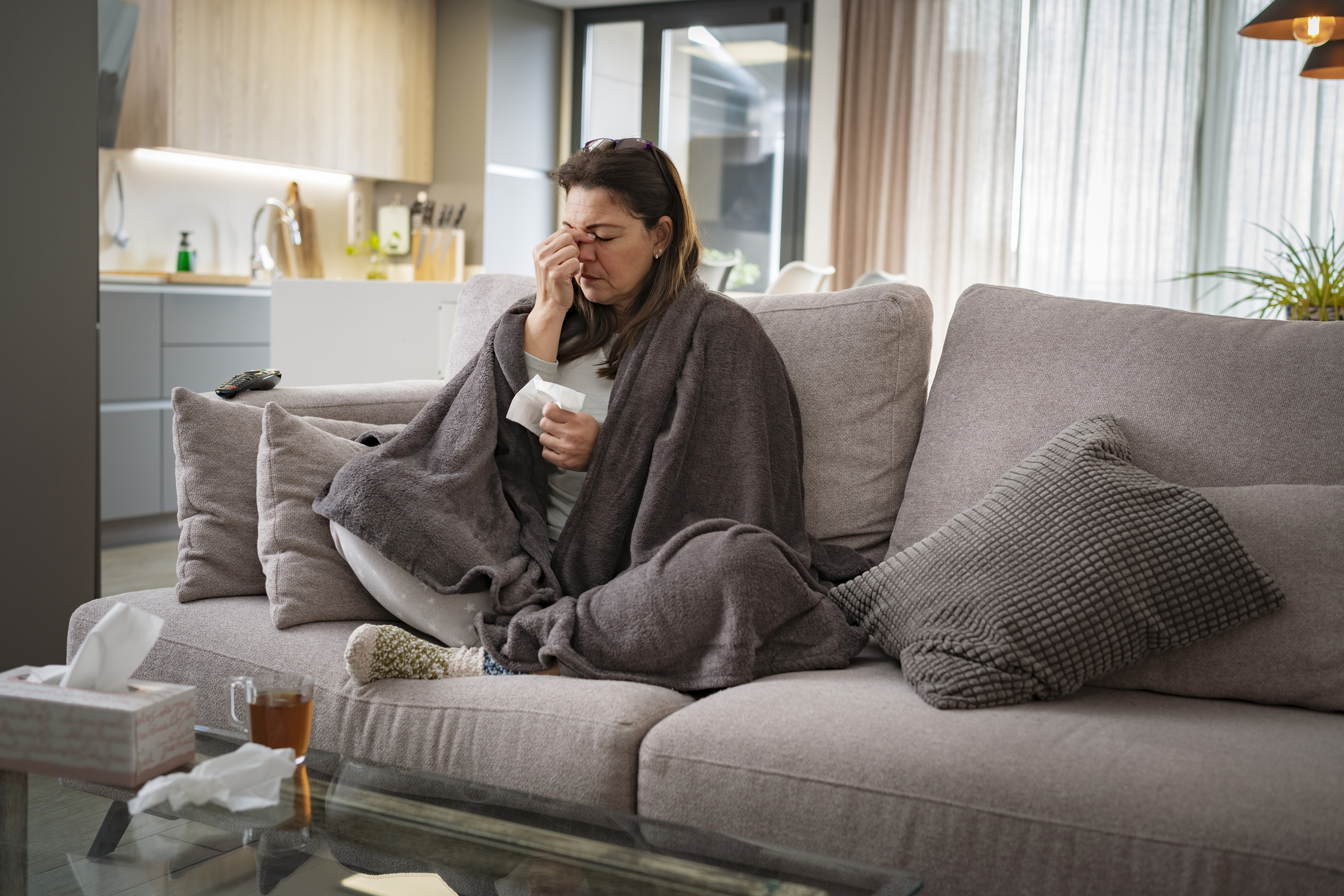 Healthcare and medicine. Woman sick with flu sitting on sofa at home massaging her nose. She is wrapped in blanket. High resolution 42Mp indoors digital capture taken with SONY A7rII and Zeiss Batis 40mm F2.0 CF lens